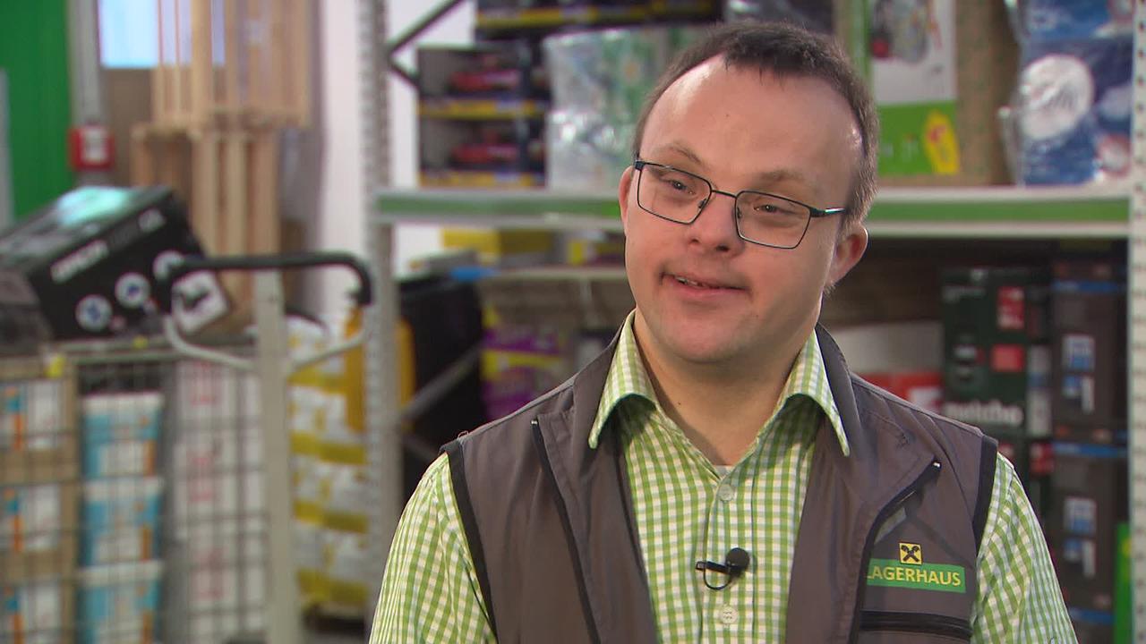Am Bild ist ein junger Mann mit Trisomie 21 in seiner Arbeitskleidung im Lagerhaus.