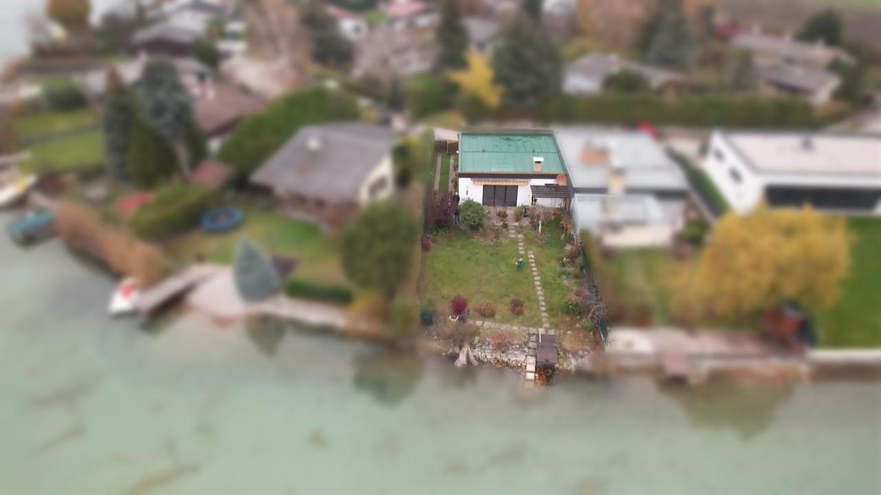 Am Bild ist eine Luftaufnahme vom Haus und dem Grundstück von Herrn J. Sein Haus liegt an einem See und steht Mauer an Mauer mit dem Nachbarhaus.