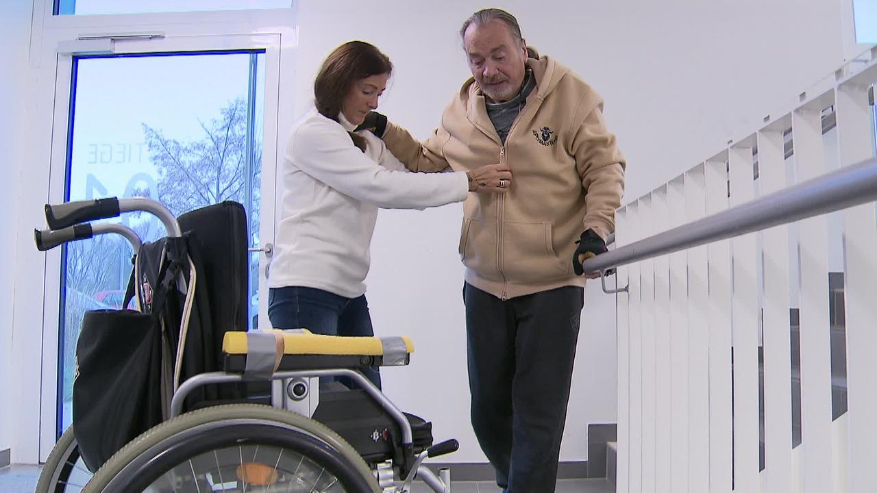 Am Bild ist Herr S. und seine Partnerin . Er wird von ihr gestützt und hält sich an einem Stiegengeländer an, um zu seinem Rollstuhl zu kommen.