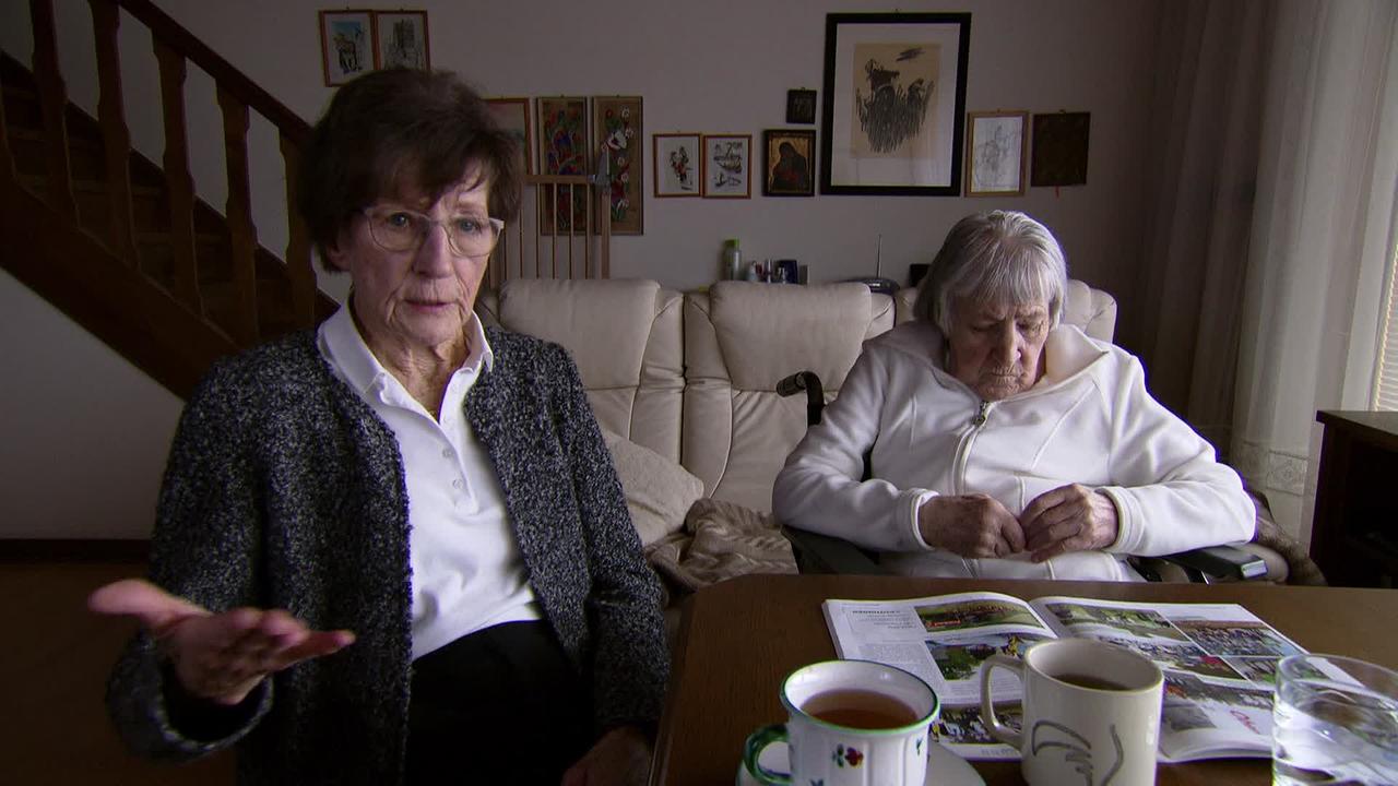 Am Bild sind zwei ältere Damen, die an einem Tisch in ihrem Wohnzimmer sitzen. Die jüngere Schwester erzählt von den Vorfällen in der Bank.