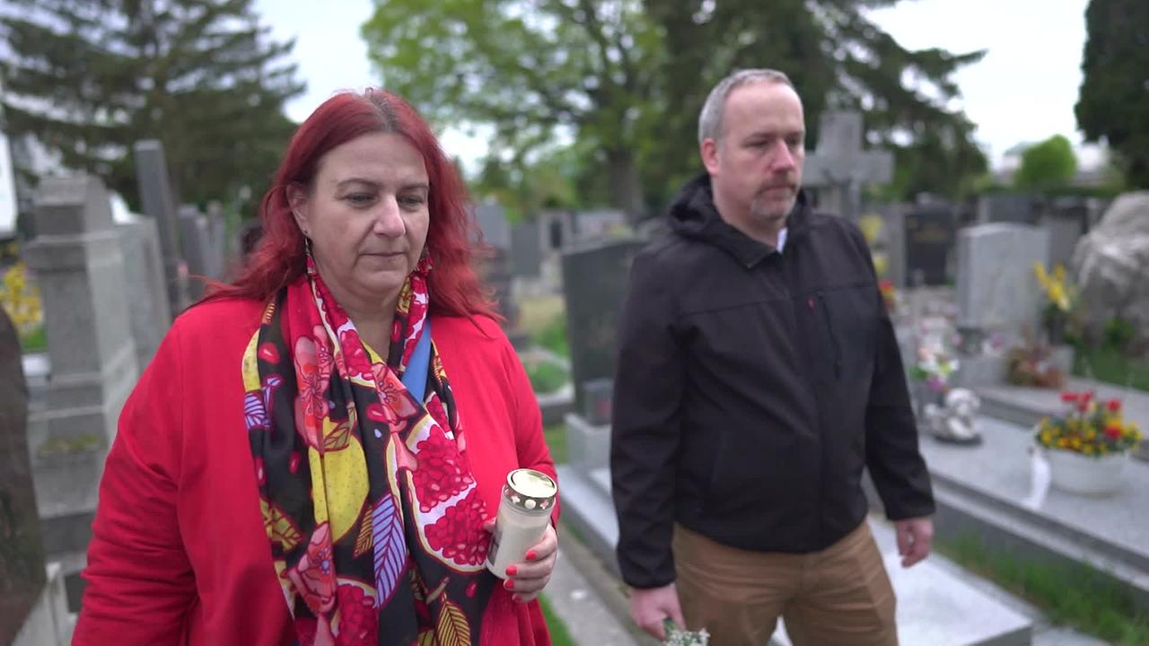 Am Bild sind die Nichte und der Neffe des Verstorbenen zu sehen. Sie stehen am Friedhof mit Blumen und einer Kerze in der Hand vor dem Familiengrab.