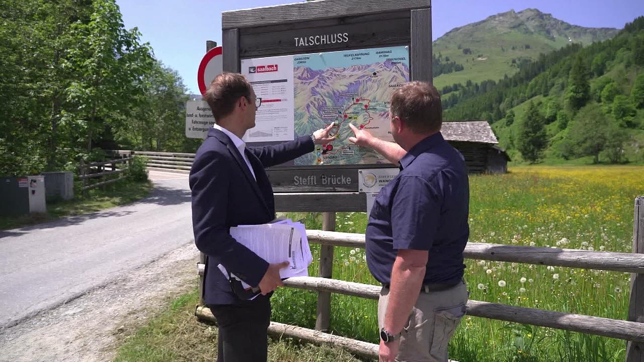 Am Bild stehen Herr W. und sein Anwalt vor einer Tafel mit der Karte vom Talschlussweg in Saalbach-Hinterglemm. Sie zeigen auf den Punkt an dem sich der Hochseilgarten befindet, in dem Herr W. einen Unfall hatte. Im Hintergrund sieht man Berge und grüne Wiesen und eine Zufahrtsstraße. 