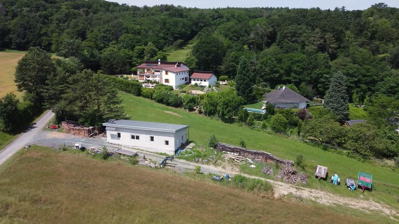 © ORF / Marco Tondolo Am Bild ist ein gemauertes Haus mit Fenstern. Links vor und hinter dem Haus sind Holzstapel und ein paar Anhänger sind geparkt. Im Hintergrund sind zwei Wohnhäuser zu sehen. Rundherum sind Wisen und Wald.