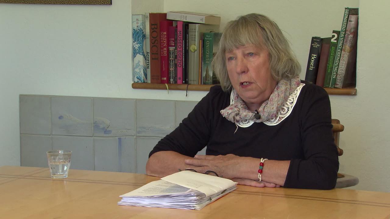 © ORF / Klaus Ibitz Am Bild ist Frau Z. Sie sitzt am Esstisch, hat Unterlagen von ihrem Krankenhausaufenthalt vor sich liegen und spricht über die Kosten ihres Aufenthaltes. Die Pensionistin hat einen dunklen Pullover mit weißem Spitzenkragen an und trägt ein farblich passendes Halstuch.