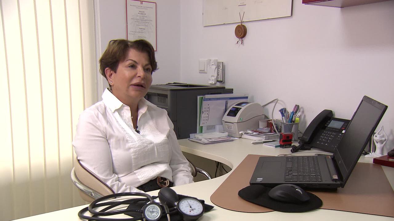Am Bild ist Frau Z., ehemalige Leiterin des Ärztefunkdienstes, in ihrer Praxis. Am Tisch liegt ein Blutdruckmesser, daneben steht ein Laptop. Sie hat kurze braune Haare und trägt eine weiße Bluse.