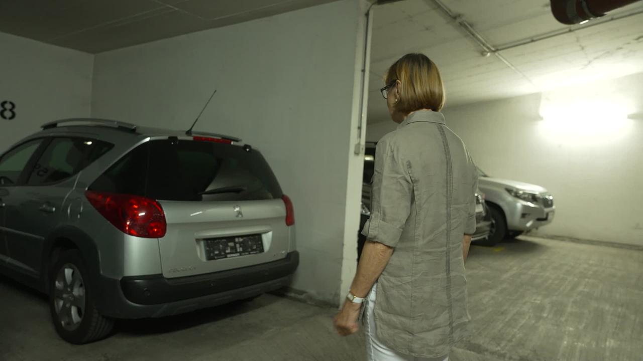 © ORF / Gerd Fellner Am Bild sieht man in einer Garage das silberne Auto von Frau B. Am Heck fehlt die Nummerntafel. Frau B. steht davor und sieht auf die fehlende Nummerntafel.
