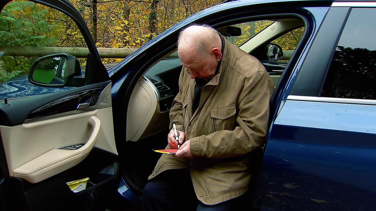 Ein älterer Mann sitzt bei offener Wagentür in seinem Auto und schreibt einen Parkschein.