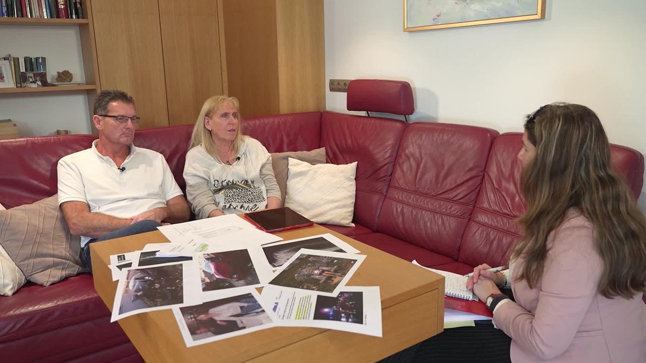 © ORF / Gabi Hanke Frau M. sitzt mit ihrem Mann auf einer roten Ledercouch in ihrem Wohnzimmer. Am Tisch vor ihnen sind diverse Fotos von ihrem Unfall. Katharina Windbichler führt ein Interview mit ihr.