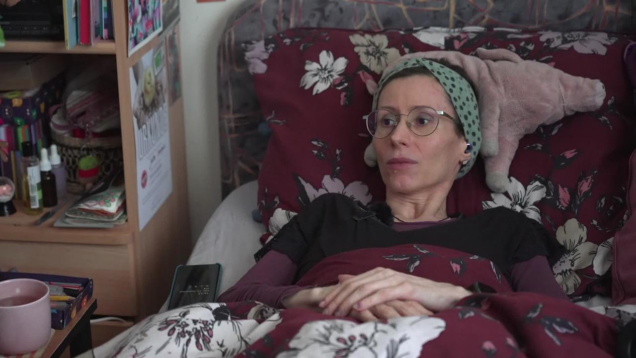 Frau S. ist eine sehr schlanke, 42jährige Frau mit Brille. Sie liegt im Bett ihres Zimmers. neben ihrem Bett sind Medikamente in einem Regal, Bücher und ein Heferl mit Getränk zu sehen.