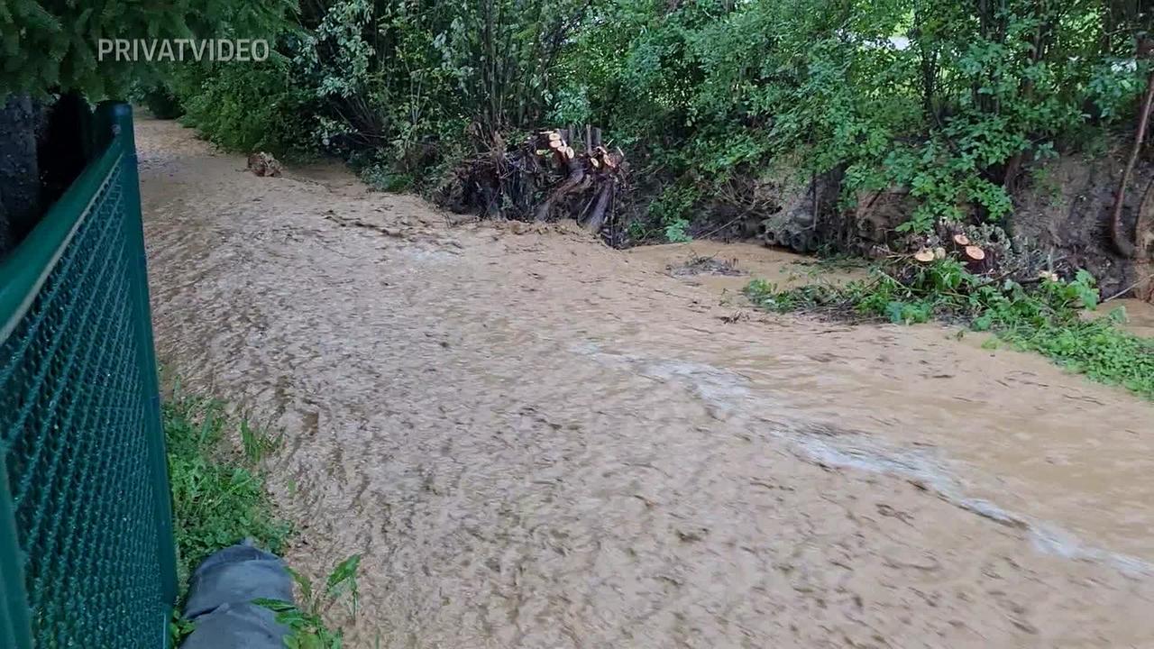 Am Bild ist ein Weg zu sehen, der komplett überflutet ist und mit Schlammmassen bedeckt ist. Auf der einen Seite ist der eingezäunte Grund von Hrn. E. auf der anderen Seite sind Büsche entlang einer Straße.