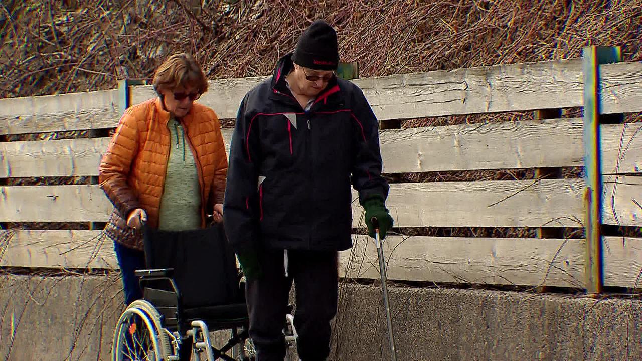 Herr T. geht mit einem Stock in kleinen Schritten an einem Zaun entlang. Seine Lebensgefährtin fährt hinter ihm mit dem Rollstuhl, falls er nicht mehr weiter kann.