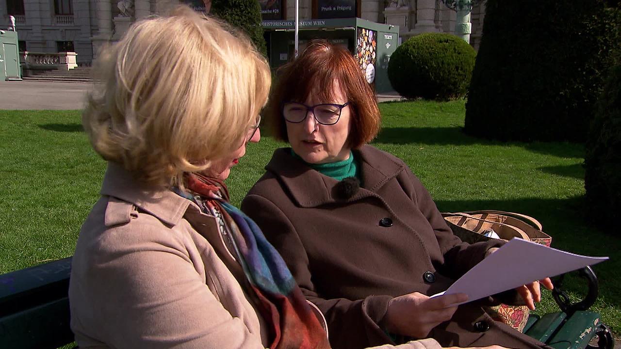 Zwei Damen, eine mit blonden Haaren, eine mit braunen Haaren, sitzen auf einer Parkbank und unterhalten sich. Eine Dame hält Papiere in der Hand.