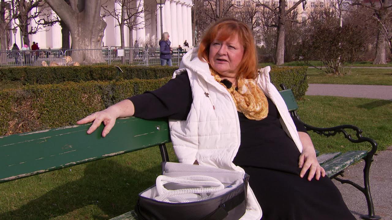Am Bild sitzt eine Dame mit rotblonden Haaren, einem schwarzen Kleid und einem weißen Daunengilet auf einer Parkbank. Neben ihr steht eine offene Tasche mit einem Beatmungsgerät.