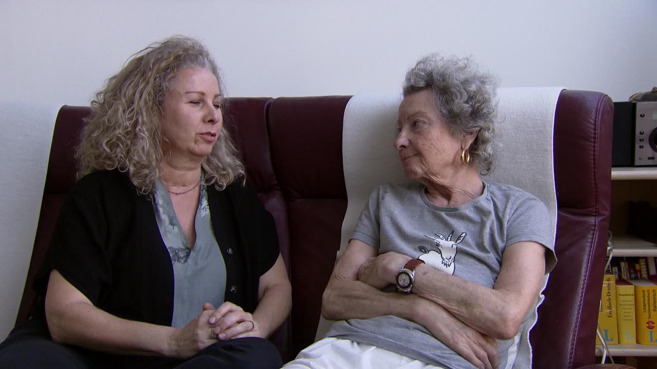 Am Bild sind zwei Frauen, die auf einer Couch sitzen. Rechts sitzt die über 70jährige Mutter mit grauen Haaren. Sie sieht ihre Tochter an.