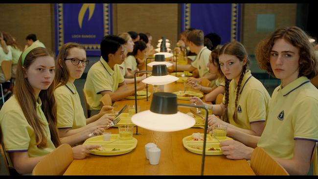 Elsa, Helen, Ragna und Fred in gelben Uniformen sitzen beim Essen an einem Tisch in einer großen Kantine, im Hintergrund sind weitere Schüler:innen an Tischen.