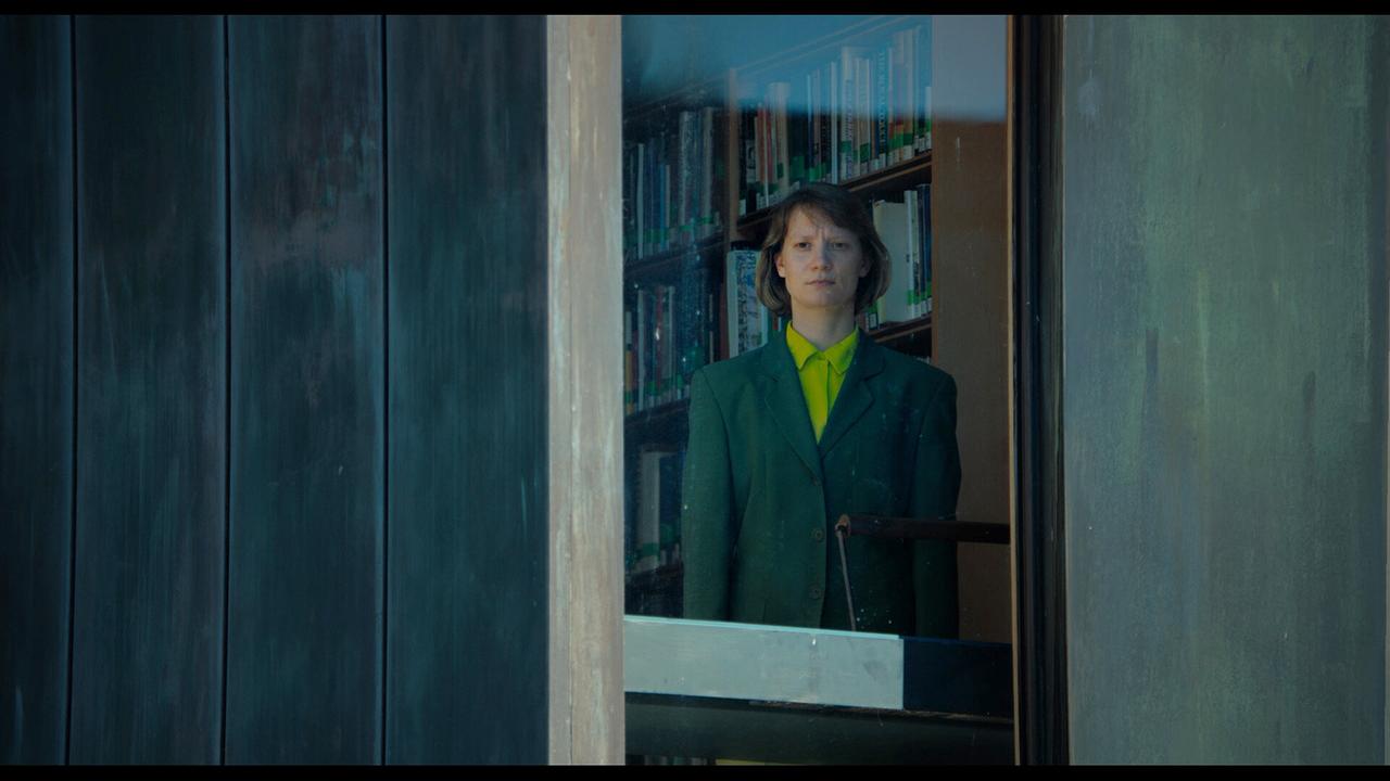 MS Novak steht hinter einer Glasscheibe vor Bücherregalen und blickt nach draußen.