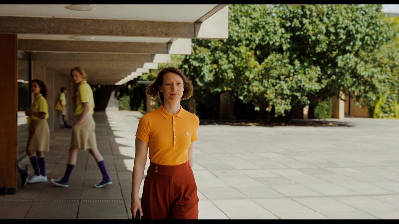MS Novak in einem orangefarbenen Polo geht allein unter einem überdachten Außenbereich mit Schüler:innen im Hintergrund.