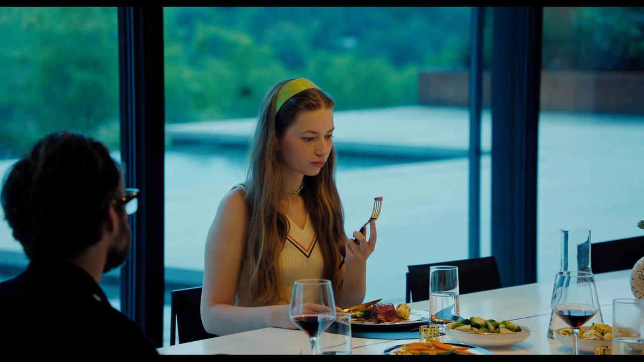 Elsa sitzt mit ihrem Vater bei Tisch in einem modernen Haus und begutachtet den Bissen, den sie auf ihrer Gabel in der Hand hat.