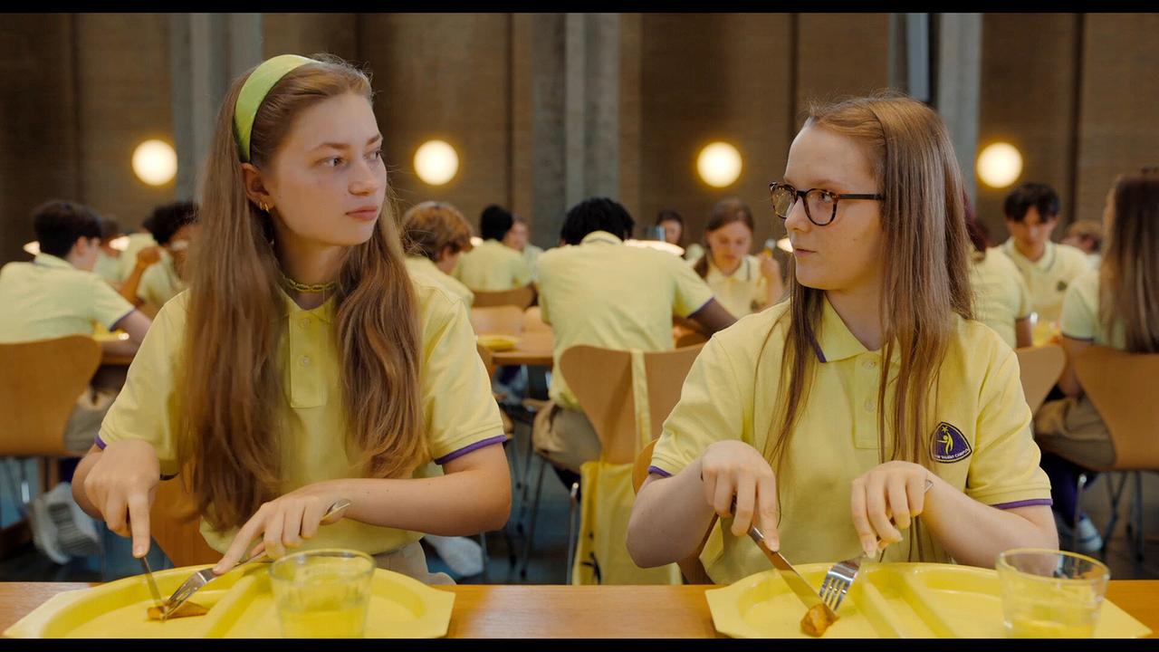 Elsa und Helen in gelben Uniformen sitzen nebeneinander und blicken sich an beim Essen in einer großen Kantine.