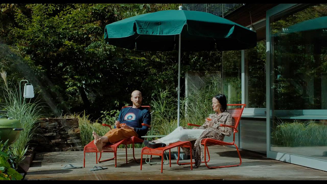 Ragnas Eltern liegen auf Liegestühlen auf ihrer Terrasse unter einem Sonnenschirm.