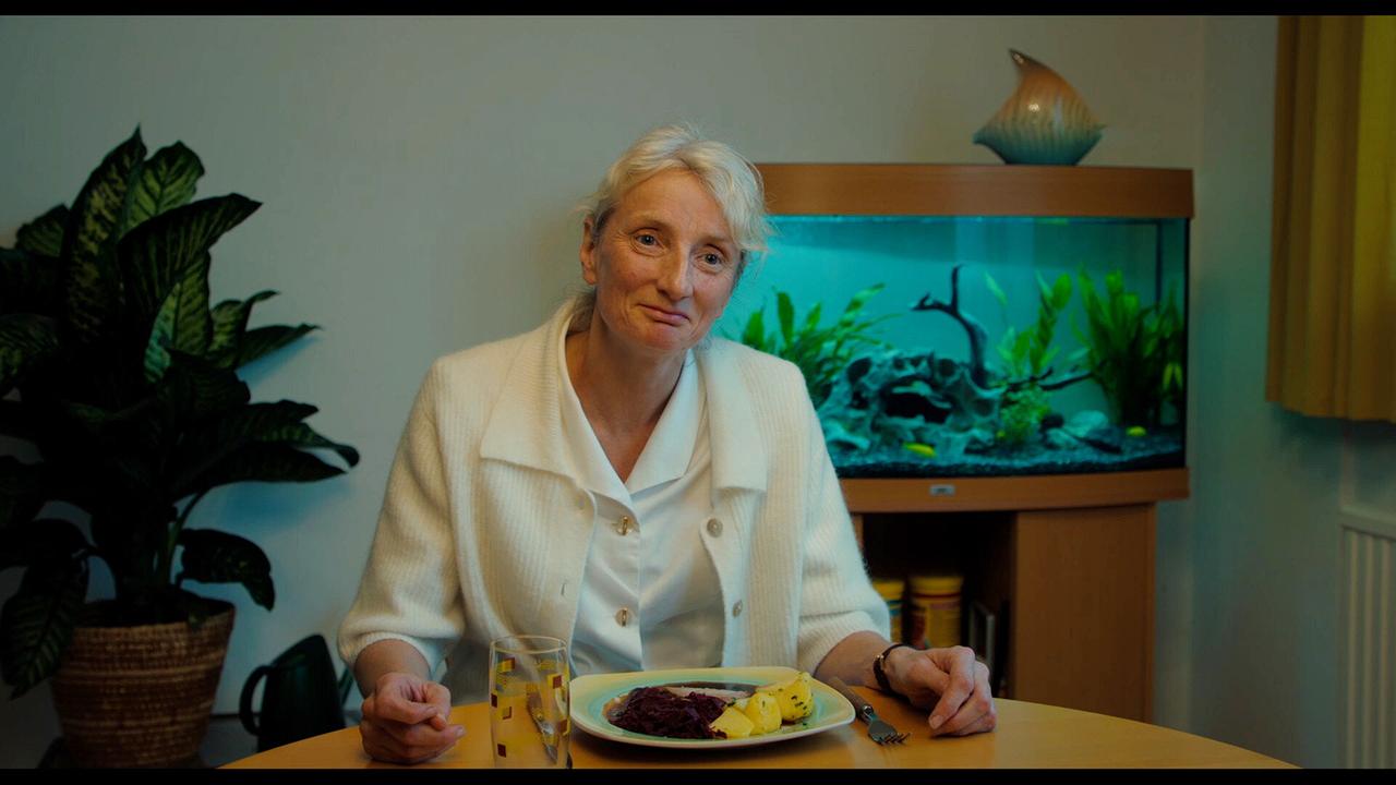 Bens Mutter sitzt am Esstisch vor einem Teller mit Essen, hinter ihr leuchtet ein Aquarium.