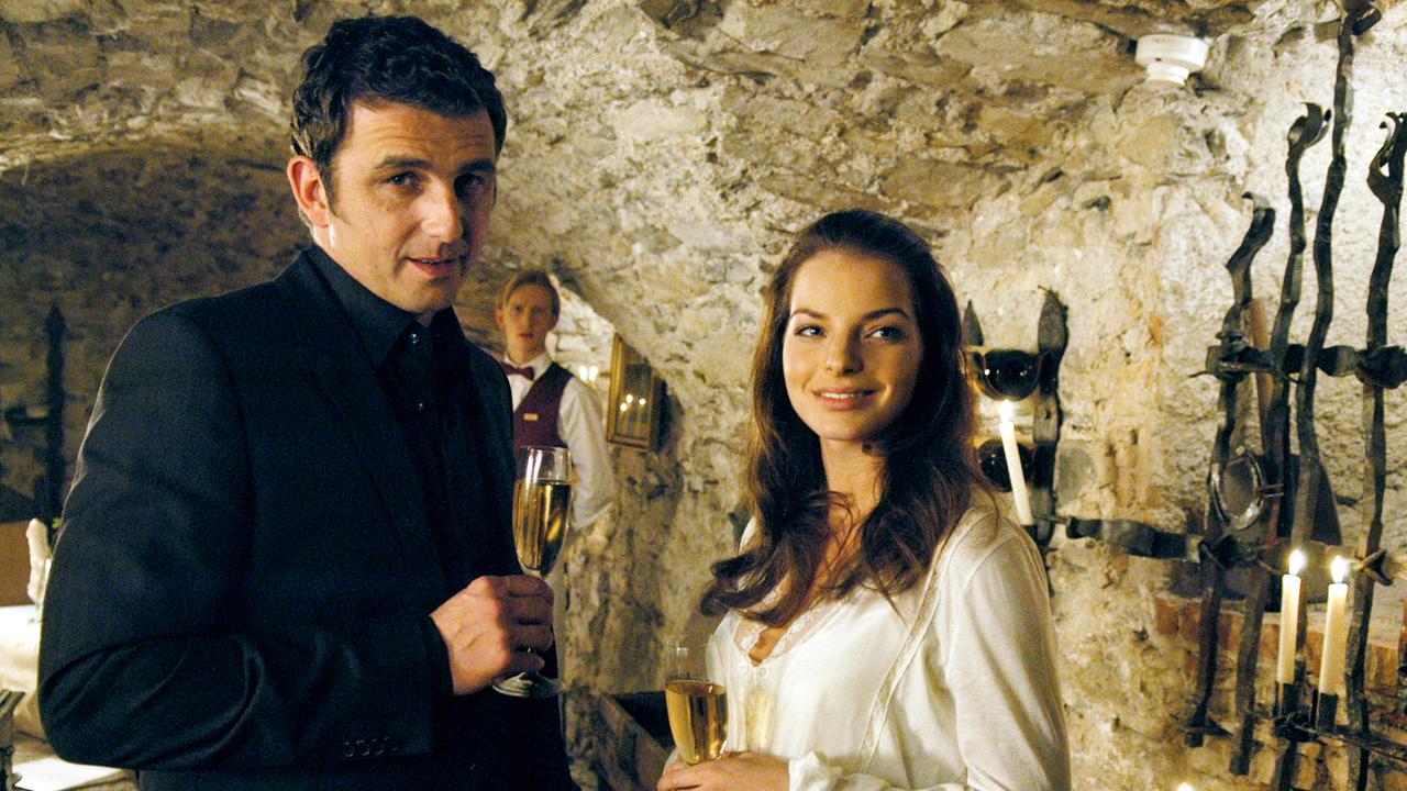 Ein Mann im schwarzen Anzug und schwarzem Hemd und eine Frau mit weißer Bluse stehen im Kellergewölbe eines Lokals, beide halten Sektflöten in der Hand und lächeln