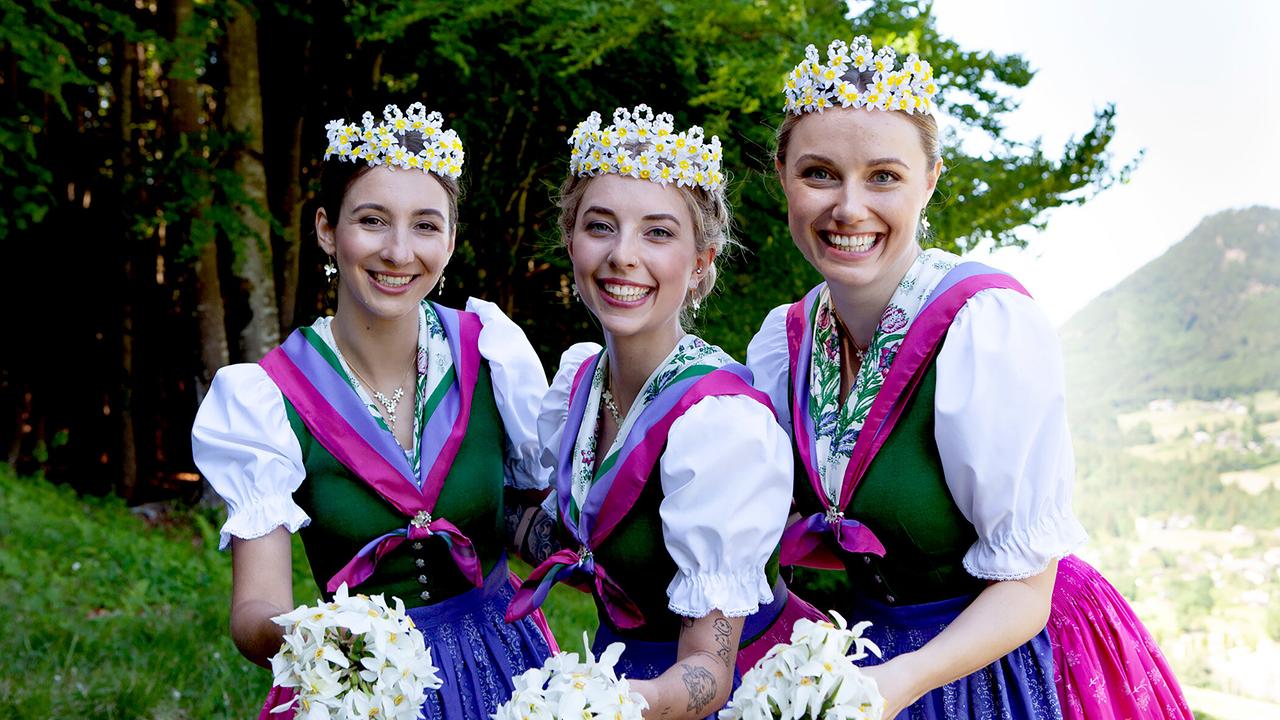 Die Narzissenhoheiten, drei Frauen in farbenfrohen Trachten mit Blumenkränzen und Blumensträußen stehen gemeinsam auf einer grünen Wiese vor einer Wald- und Berglandschaft.