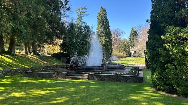 Giardini Botanici di Villa Taranto mit einem zentralen Springbrunnen, umgeben von sattgrünem Rasen, hohen Bäumen und klarem, blauem Himmel.