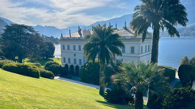 Blick auf die Villa Melzi d'Eril bei Sonnenschein umgeben von Palmen, gepflegtem Rasen und Comer See mit Bergkulisse im Hintergrund.