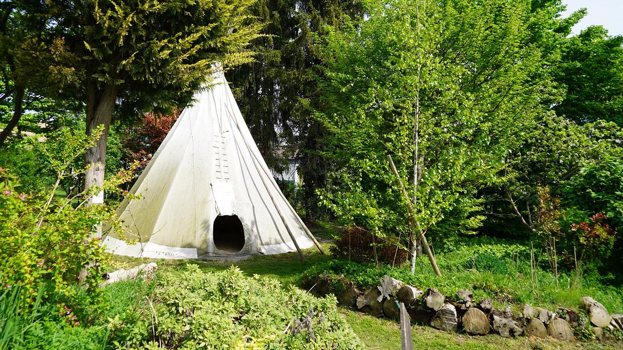© Leopold Mayrhofer/Natur im Garten Ein weißes Zelt steht in einem grünen Schaugarten umgeben von Bäumen und Sträuchern und einem kleinen Steinhaufen.