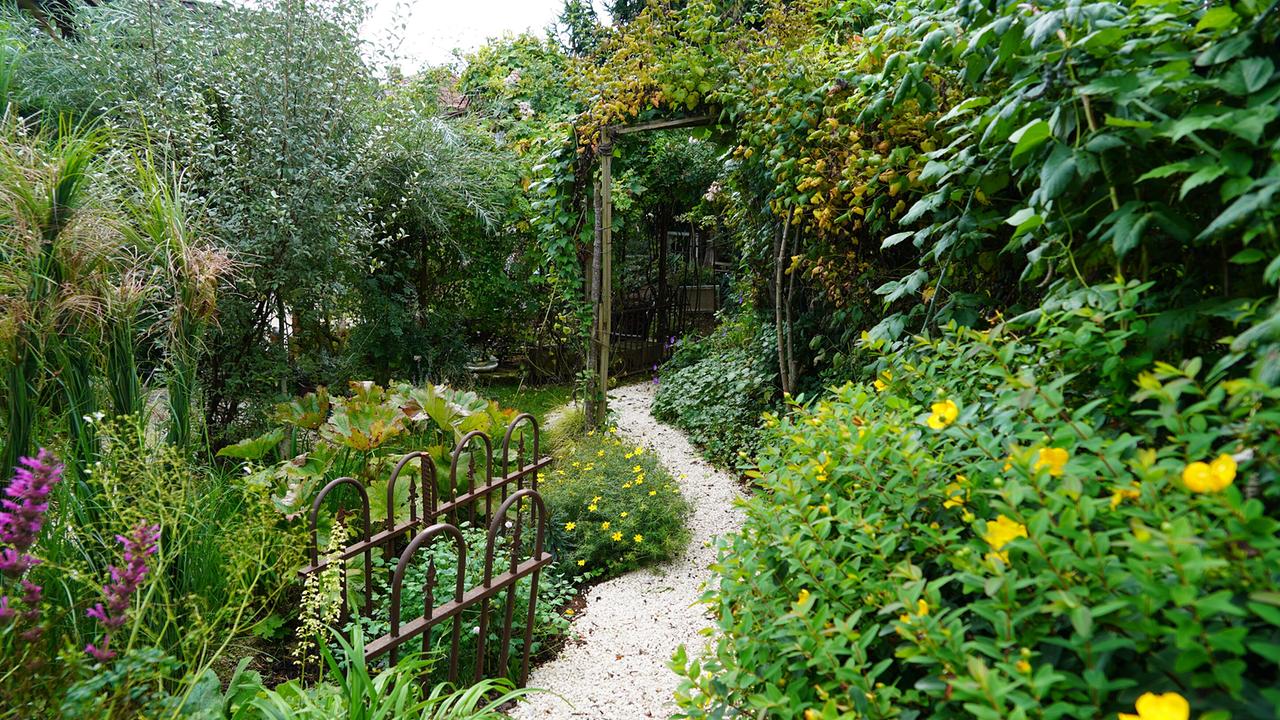 © Leopold Mayrhofer/Natur im Garten/Leopold Mayrhofer, A.Wildgansstr. 3382 LOOSD. Ein geschwungener Kiesweg führt durch einen üppig bepflanzten Garten mit Hügeln und einem kleinen Torbogen.