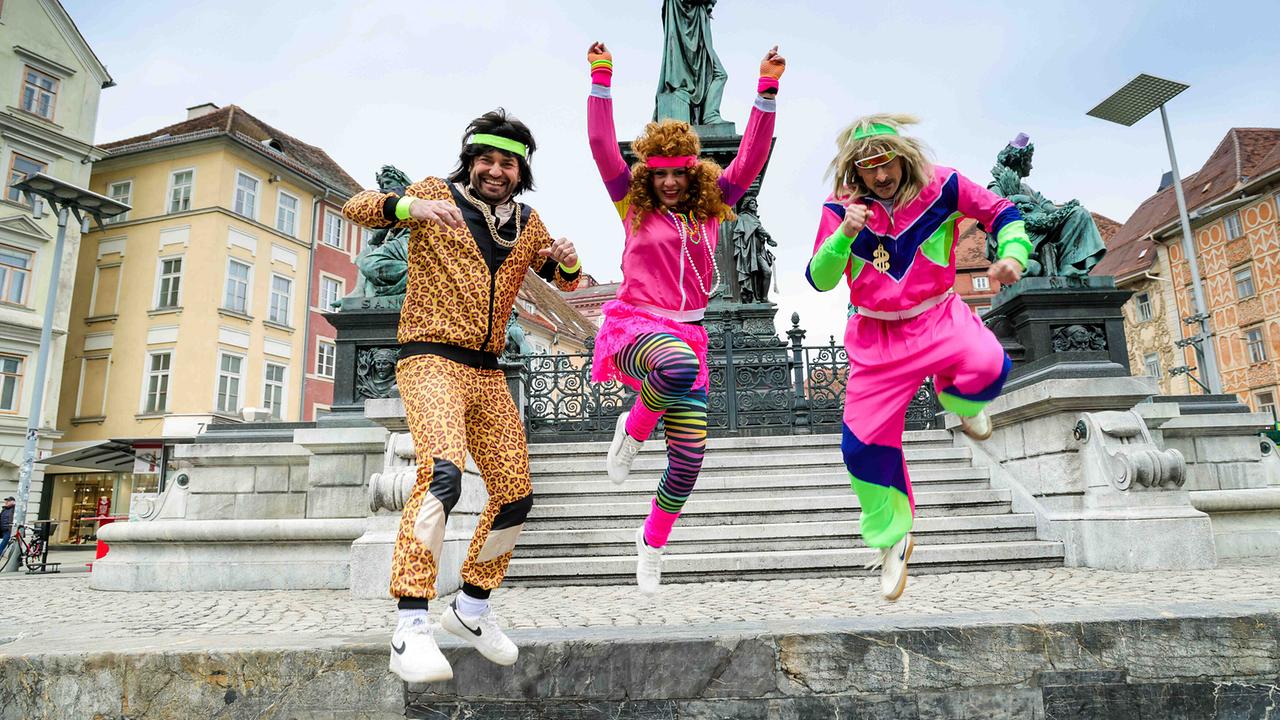 © ORF/Kleine Zeitung/Stefan Pajman Thomas Seidl, Kathi Wenusch und Florian Prates in bunten 80er‑Jahre‑Kostümen springen ausgelassen vor den Stufen zu einem Denkmal in der Grazer Innenstadt.