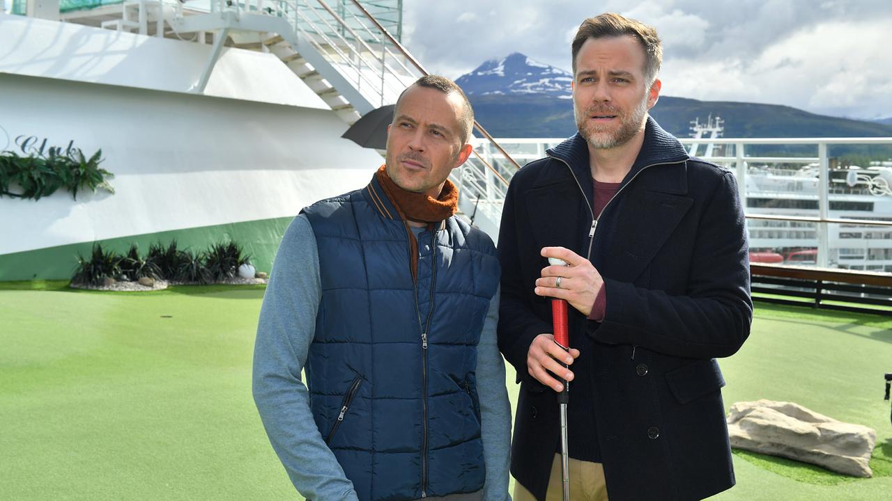 Nikolaus Andresen und Dominik Dahlke stehen auf dem Deck des Traumschiffes bei einer Minigolfbahn, einer hält einen Golfschläger, im Hintergrund sind Berge zu sehen.