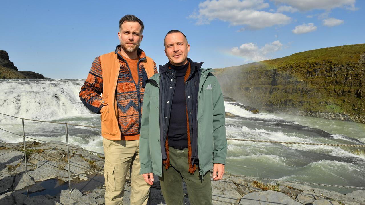 Nikolaus Andresen und Dominik Dahlke stehen vor einem Wasserfall in einer felsigen Landschaft in Island.
