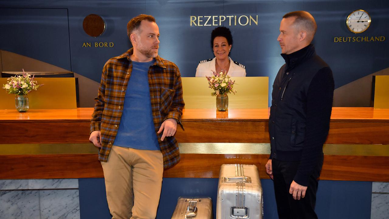 Dominik Dahlke und Nikolaus Andrese stehen an einer Rezeption mit Koffern, während Hanna Liebhold hinter dem Tresen lächelnd wartet.