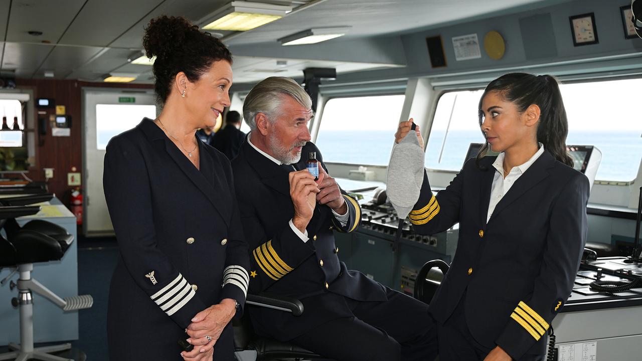 Der Staff-Kapitän Martin Grimm gibt Hanna Liebhold und Dr. Jessica Delgado in Uniform auf der Brücke eines Schiffes Tipps für ihre Übernachtung in Islands Wäldern und zeigt ein kleines Fläschchen, während Dr. Delago eine Schlafmaske in Händen hält.