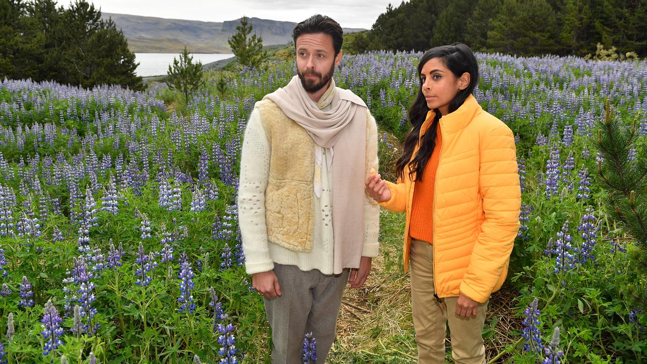 Dr. Jessica Delgado ist vom Weg abgekommen und erhält Unterstützung von dem geheimnisvollen Jon. Sie stehen in einem blühenden Feld mit violetten Blumen, umgeben von Bäumen und sanften Hügeln.