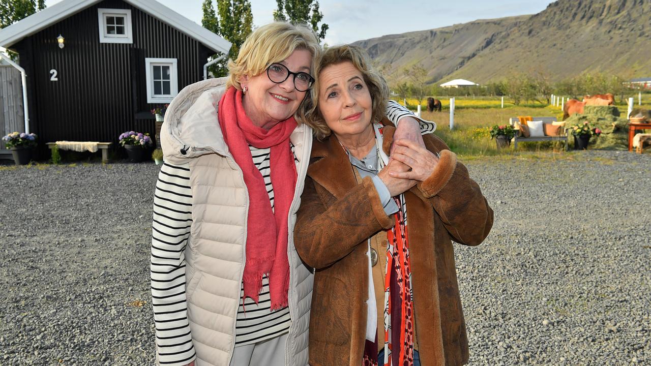 Evelyn Küpper und Toni Meissner umarmen sich freundschaftlich auf einem Hof vor einem kleinen Haus, Bergen und Weideland im Hintergrund.