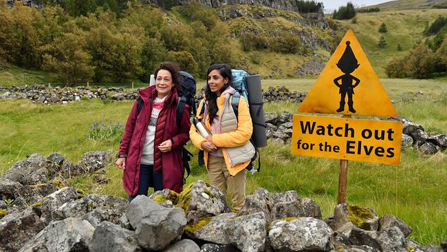 Gemeinsam erkunden Hanna Liebhold und Dr. Jessica Delgado (Collien Fernandes, r.) die spektakuläre Landschaft Islands und stehen mit großen Rucksäcken in einer grünen, felsigen Landschaft neben einem gelben Warnschild mit der Aufschrift „Watch out for the Elves“.