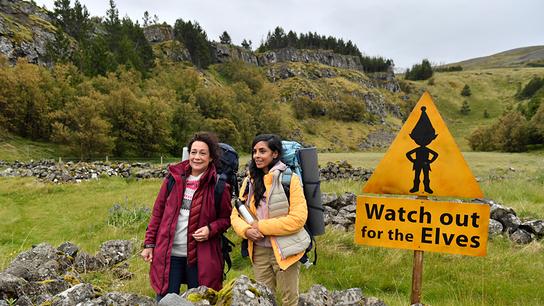 © ORF/ZDF/Dirk Bartling Gemeinsam erkunden Hanna Liebhold und Dr. Jessica Delgado (Collien Fernandes, r.) die spektakuläre Landschaft Islands und stehen mit großen Rucksäcken in einer grünen, felsigen Landschaft neben einem gelben Warnschild mit der Aufschrift „Watch out for the Elves“.