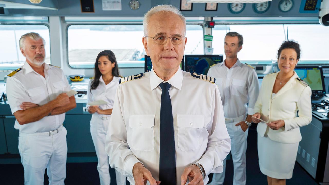 Harald Schmidt als Kreuzfahrt-Direktor in der Mitte und vier weiteren Crewmitglieder im Hintergrund stehen auf der Brücke und halten Unterlagen zu teambildenden Maßnahmen in den Händen.