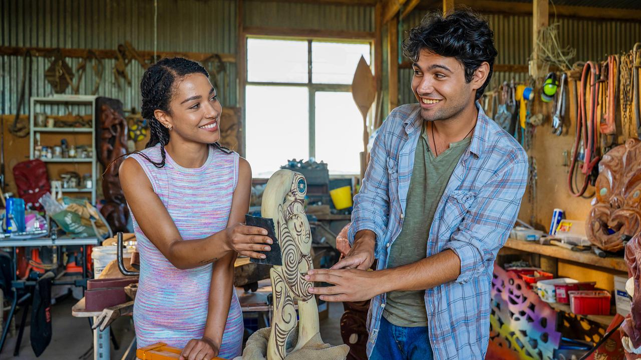 Luna Baptiste und Max Crean arbeiten gemeinsam an einer verzierten Holzskulptur in einer Werkstatt und lächeln sich dabei an.