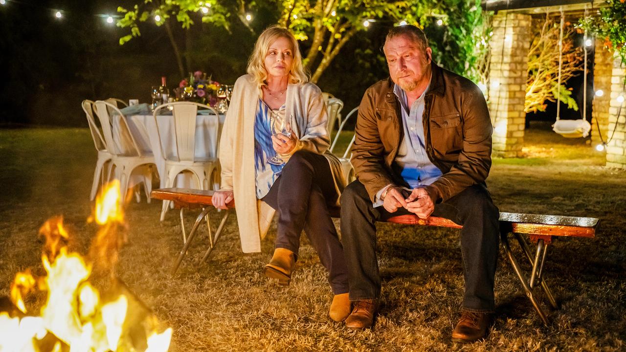 Susanna Simon und Matthias Komm sitzen am Lagerfeuer auf einer Bank, während im Hintergrund eine festlich gedeckte Tafel steht.