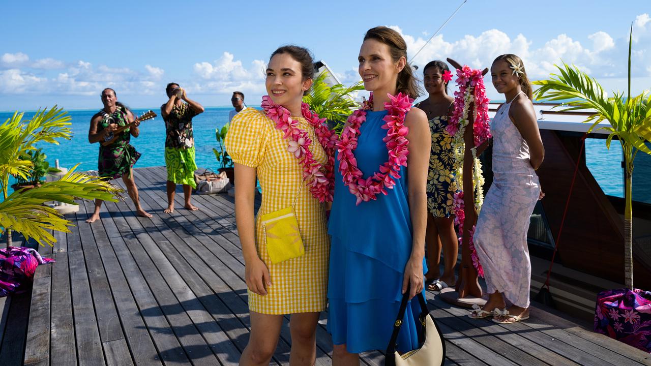 Elsa Westrup und ihre Tochter Nora mit Blumenketten um den Hals stehen auf einem Holzsteg mit Palmendeko, während im Hintergrund Musiker spielen.