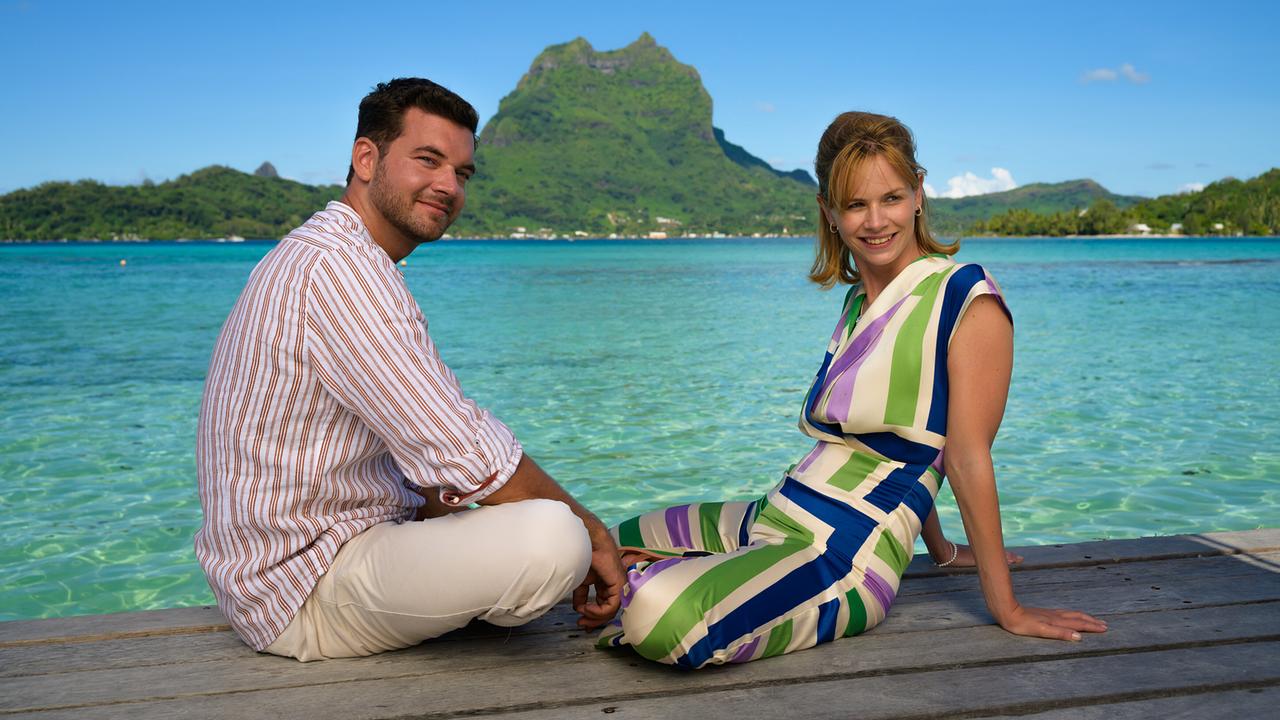 Toni Wittig sitzt mit Landschafsgärtner Basti Kugler entspannt auf einem Steg mit Blick auf türkisfarbenes Wasser und eine grüne Insel.