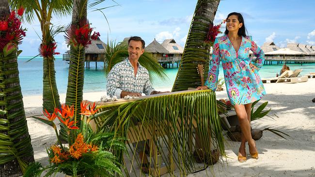 Dr. Jessica Delgado steht neben Kapitän Max Parger, der ein Musikinstrument an einem paradiesischen Strand spielt, umgeben von Palmen und Blumen und türkisfarbenem Meer.