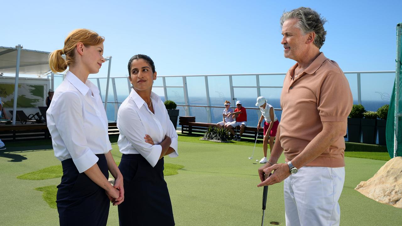 Toni Wittig, Amrei Seibold und Ulrich von Stein unterhalten sich auf einem Minigolfplatz an Deck eines Kreuzfahrtschiffs.