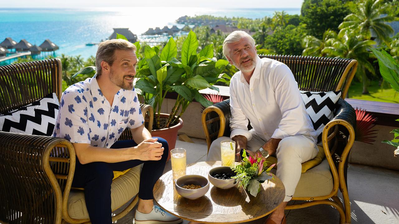 Paul Richter und Martin Grimm sitzen entspannt bei Getränken auf einer Terrasse mit Meerblick.