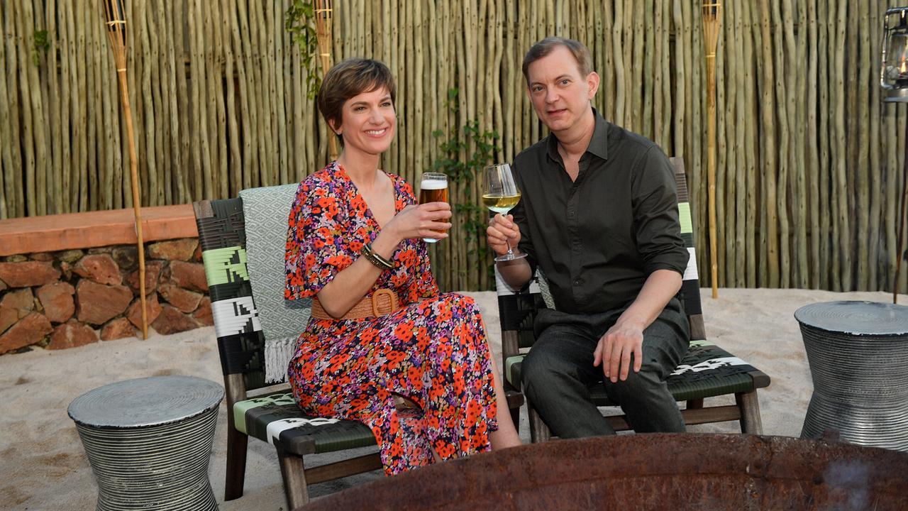 Katja Sommer und Björn Wagner stößen abends mit Wein und Bier im beleuchteten Outdoor‑Bereich einer Lodge an.