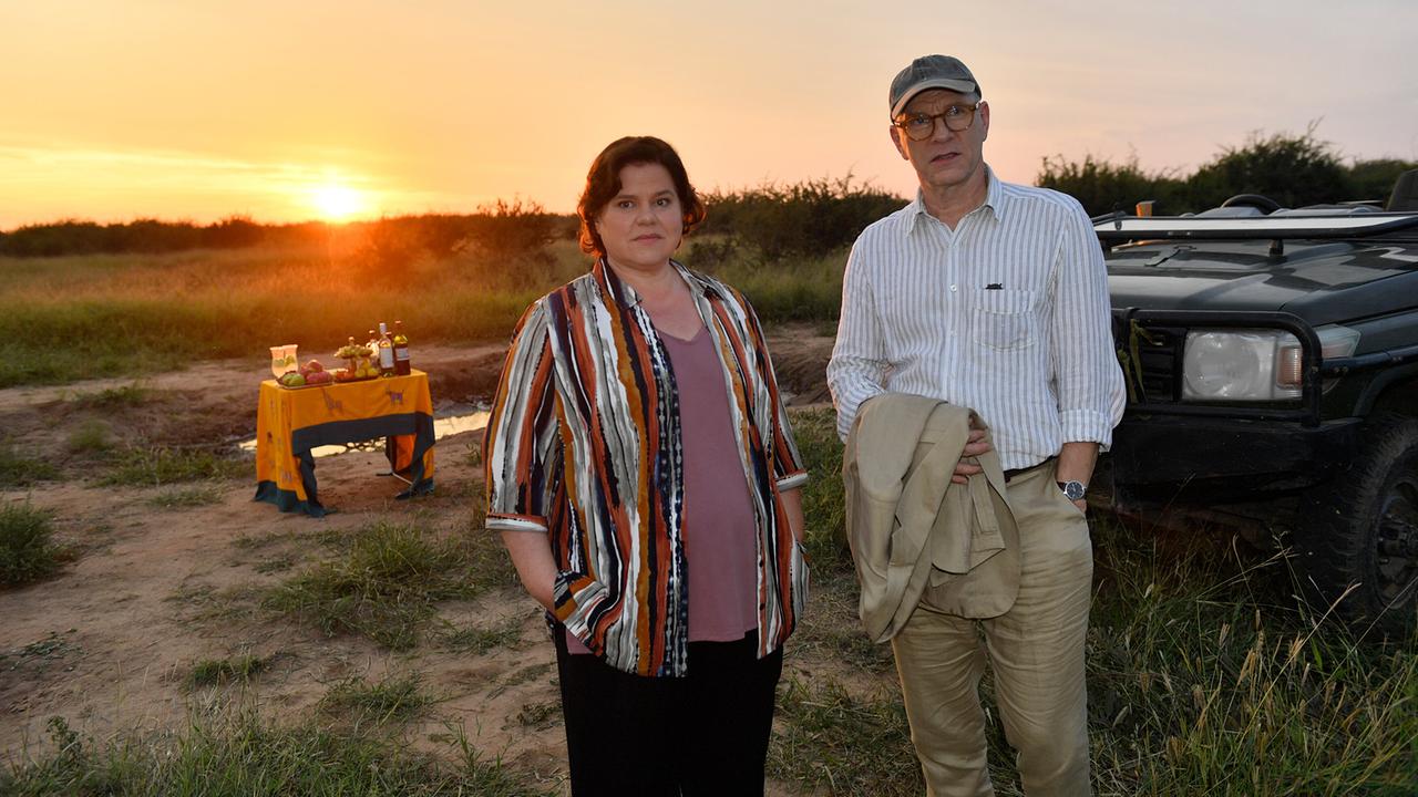 Emilia Gronewald und Oliver Stiller posieren vor einem Safari‑Jeep in freier Natur bei Sonnenuntergang neben einem kleinen Getränketisch.